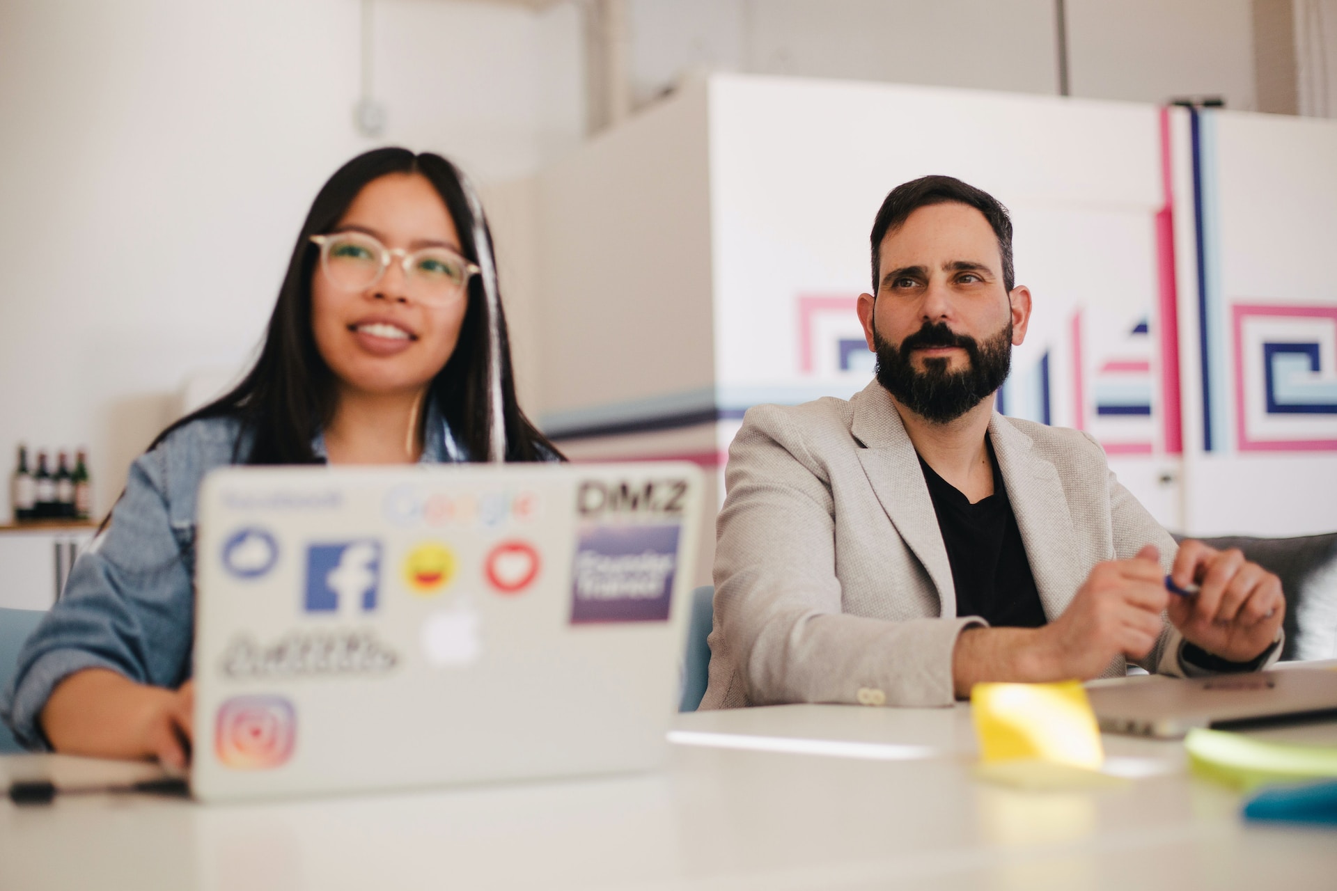 two people--one with long hair and glasses, one with a beard and beige suit--sitting at a table with a laptop looking into the distance